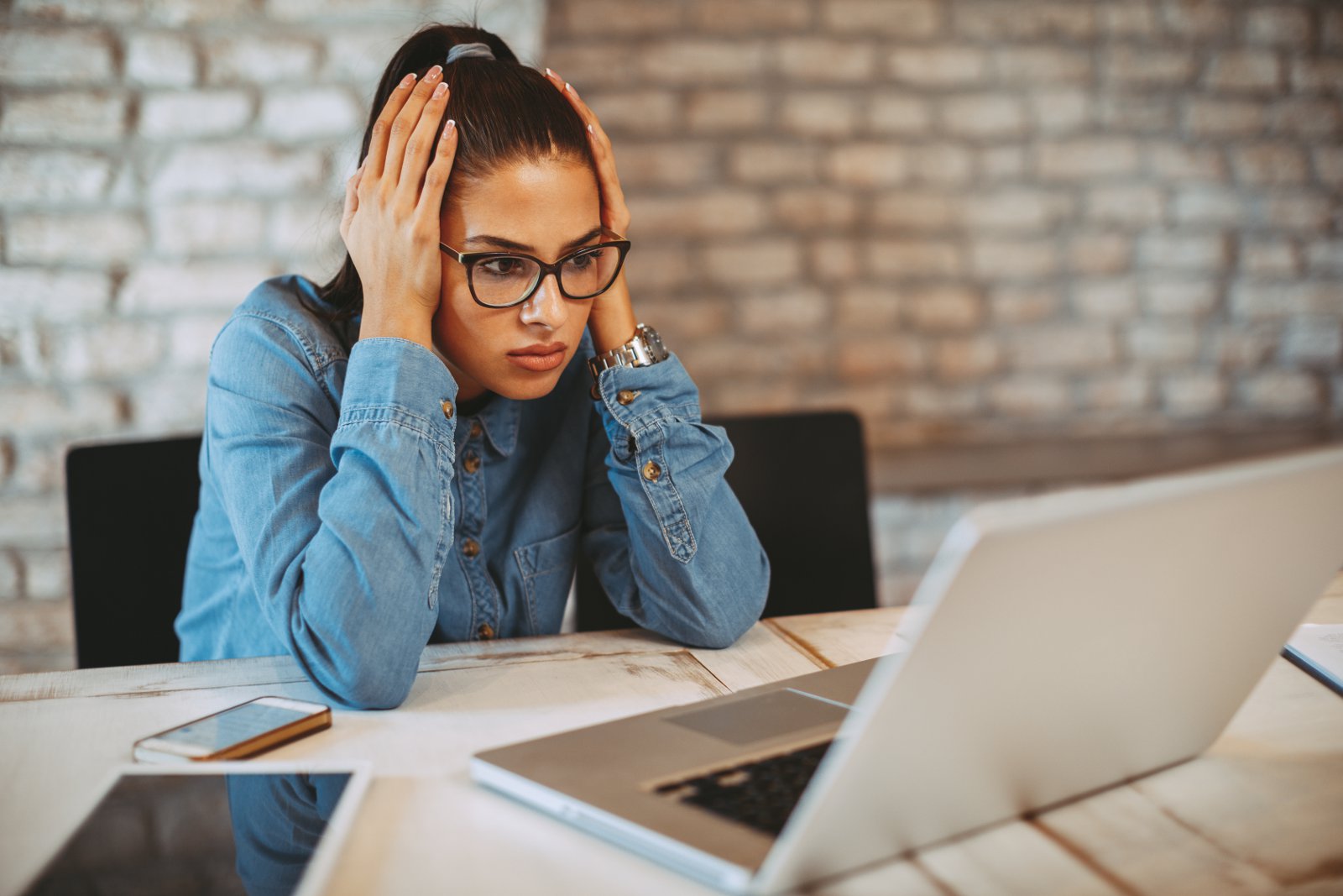 Employee sitting at desk looking stressed while working on a laptop in a busy office